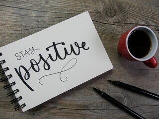 STAY POSITIVE lettering in notebook on wooden surface with coffee and pens