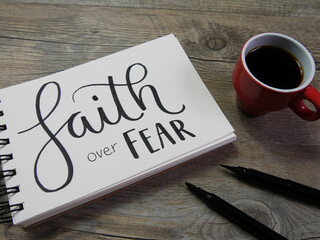 FAITH OVER FEAR lettering in notebook on wooden surface with coffee and pens
