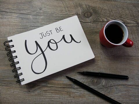 JUST BE YOU lettering in notebook with cup of espresso and pens on wooden desk