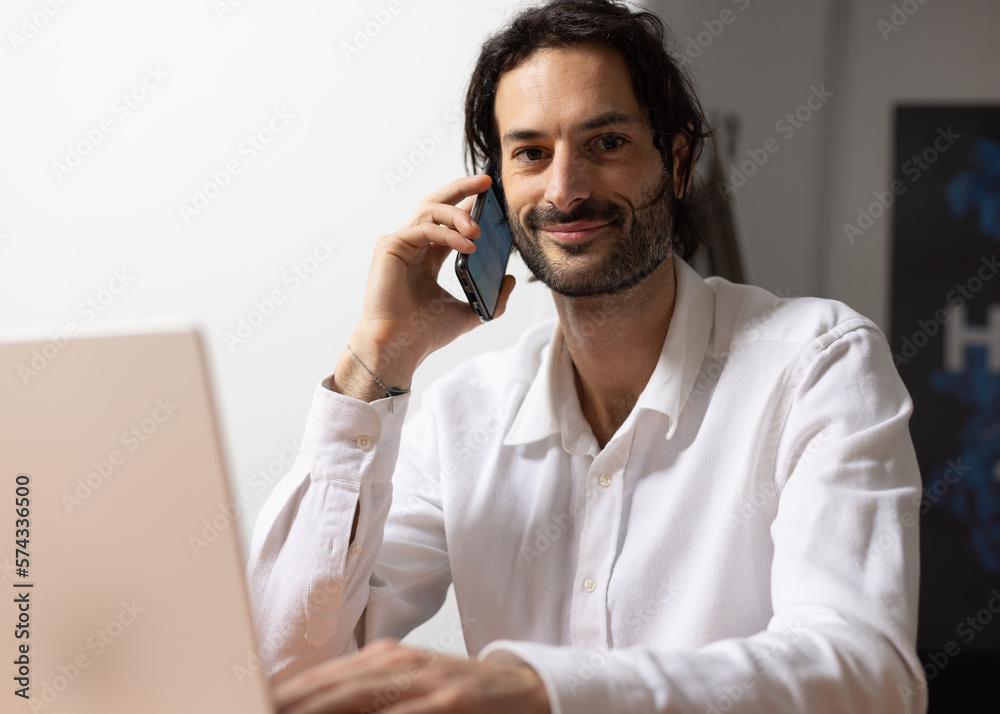 Sticker portrait d'un jeune homme souriant, employé de bureau assis devant son ordinateur au bureau. Il téléphone avec son smartphone