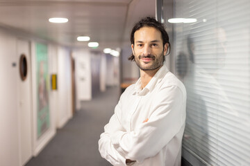 Portrait d'un beau jeune homme de 30 ans, debout, les bras croisé, dans le couloir de son bureau. Il est serein et souriant, heureux au travail. Symbole de la réussite.