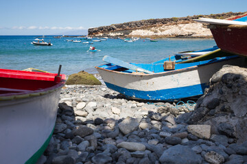 Fototapeta na wymiar Cheap boats on a pebble beach. Red and white boat on the left, blue and white boat on the right. Bay of turquoise waters and brown and yellow hills in the background. La Caleta, Tenerife, Canary Islan