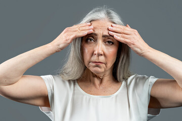 Sad old european lady with gray hair touches her face, wrinkles with hands, isolated on gray...