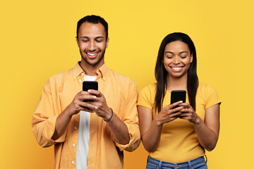 Cheerful young african american husband and wife chatting on smartphones, isolated on yellow background