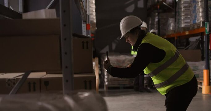 4k slow-motion video of the female warehouse manager using the mobile phone to scan the boxes while sorting the storage.  The coworkers can be seen in the background doing their work.