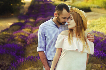 A couple is standing in a lavender field. The blonde in a pink dress and with a straw hat, against the background of a purple field of flowers. Romance in relationships, aromas.