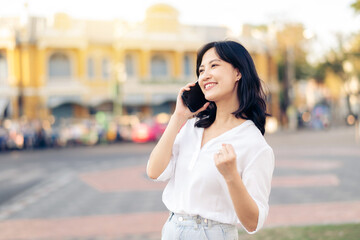 Portrait beautiful young asian woman receiving good news on mobile phone and arms raised feeling cheerful ,happiness. around outdoor street view in a summer day