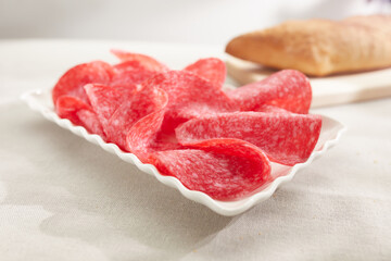 salami slices on a white plate with bread in the background. sausage close-up. meat delicacy on a sunny day. traditional mediterranean snack on a light surface. dried sausage serving. antipasta macro