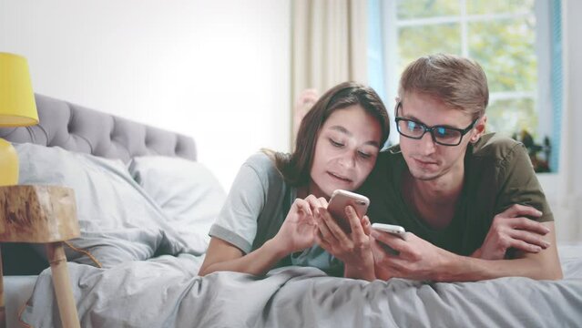 Millennial couple talking, using smartphone lying on bed together. Realtime
