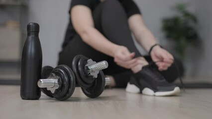 woman preparing for training at home and tying shoelaces on sneakers - dumbbells and bottle of water on first plan - Powered by Adobe