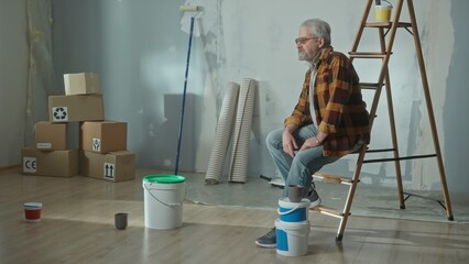 A tired elderly man sits on a stepladder while resting. A pensioner poses in a room in front of a painted wall, cardboard boxes, paint cans and wallpaper rolls. The concept of repair.