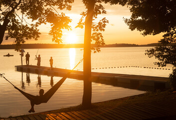 Fototapeta na wymiar Summer holiday at the lake. Setting sun, people fishing on jetty and resting on hammock, copy space.