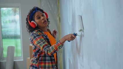 African American woman in red headphones paints the wall with white paint using paint roller and...