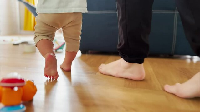 baby newborn first steps. mother teaches daughter baby to take the first steps with bare feet on the floor. baby walks barefoot on the floor at light home learning. baby newborn first steps indoors