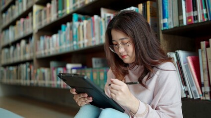 Asian woman wearing glasses is using a tablet sitting in the library