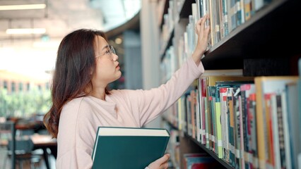 Asian woman wearing glasses is choosing books on the library bookshelf