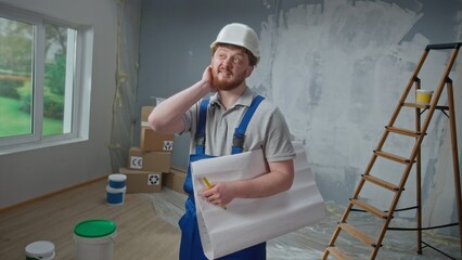 Male foreman is looking through large sheet with an apartment plan and thinking over repair project in flat. Redhead man with beard in blue construction overalls and white helmet is planning repair.