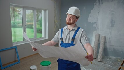 Male foreman is looking through large sheet with an apartment plan and thinking over repair project in flat. Redhead man with beard in blue construction overalls and white helmet is planning repair.