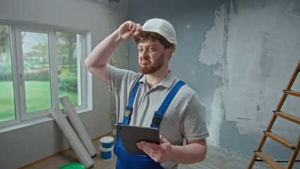 Male foreman is planning renovation project in an apartment using tablet. Redhead master man thinking, viewing and visualizing ideas against background of apartment in process of renovation.