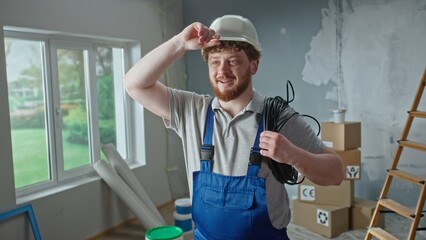 Foreman or electrician in blue overalls, white helmet and coil of black cable in hands. Redhead man is planning repairs in an apartment against backdrop of ladder, cardboard boxes and window.