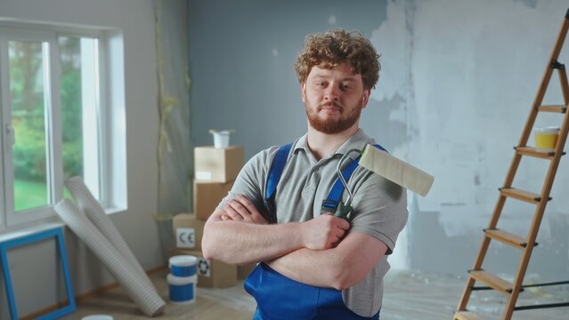 Repairman in blue overalls is looking confidently at camera and crossing arms with paint roller. Portrait of redhead man is posing against backdrop of renovation apartment, ladder, cardboard boxes