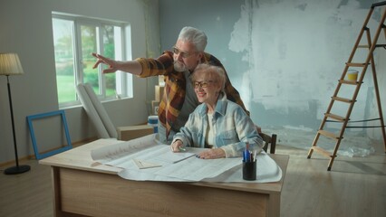 Elderly man and woman are looking through sheet with plan of an apartment and discussing renovation project. Happy aged couple is sitting at a table and planning the improvement of their home.