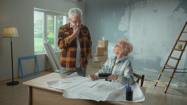 Elderly Man And Woman Are Calculating Expenses Of Repairing An Apartment On Calculator. Aged Couple Is Planning The Improvement Of Their Home. Concept Of Repair, Decoration, Interior Design.