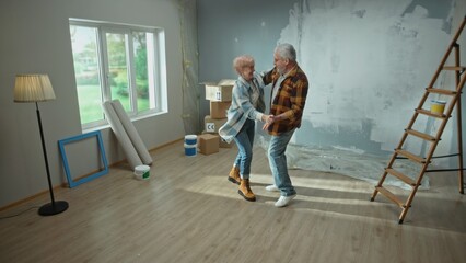Elderly man and woman dance cheerfully and smile. A couple of pensioners have fun and celebrate housewarming. Ladder, cardboard boxes and a framed window. The concept of repair.