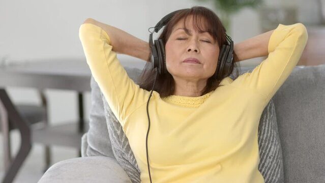 Beautiful mature woman wearing headphones relaxing listening to music laying back hands behind head eyes closed sitting in comfortable home living room sofa