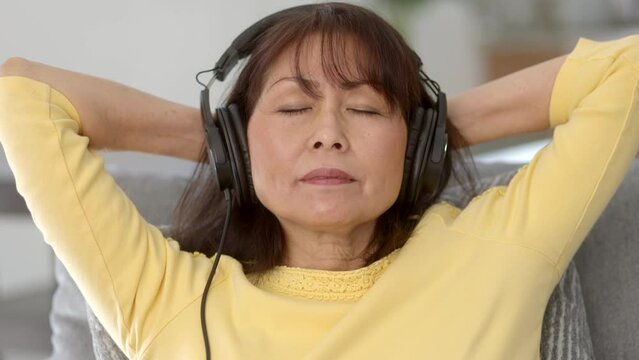 Closeup attractive middle age woman wearing headphones relaxing listening to music laying head back eyes closed sitting in comfortable home living room sofa