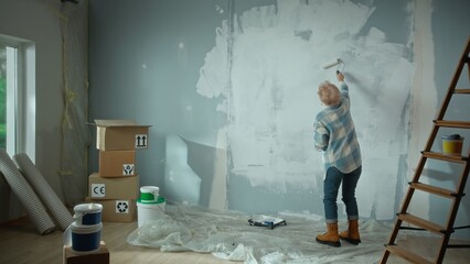 Elderly woman is painting wall with white paint using paint roller. Female pensioner is making repairs to their apartment, in background of window, stepladder, cardboard boxes, wallpaper, paint. Back