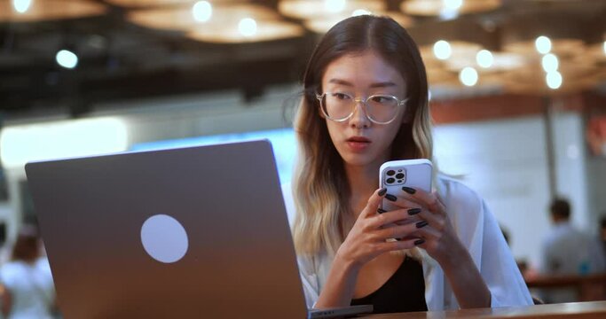 Asian young female student use mobile phone app and computer to work or study, sitting in coffee shop. Teenager woman in glasses looking at mobile phone screen and use laptop computer in stylish cafe