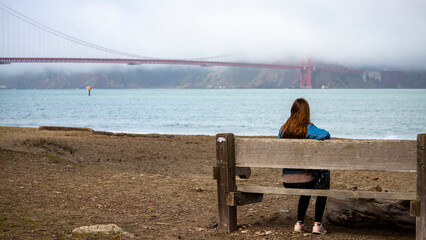 long haired girl sits on a bench on a beach and admires famous popular large Golden Gate Bridge hidden in a fog in San Francisco, California, USA. Winter in San Francisco. 