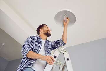 Happy man changing a light bulb at home. High angle shot of a young man in a checkered shirt standing on a step ladder and placing a modern energy-saving lightbulb in a white lamp on the ceiling - Powered by Adobe