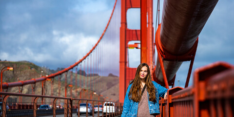 a beautiful long-haired model in a denim jacket stands on the famous golden gate bridge in san francisco during cloudy weather