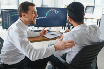 Two successful trader sitting in office and looking at screen, checking cryptocurrency information data on finance market graph, pointing on monitor