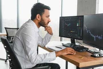 Side view of smart Indian crypto trader, investor, analyst broker, using laptop and smartphone analyzing digital cryptocurrency exchange, stock market charts, thinking of investing and funds risks.