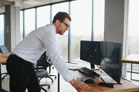 Man trader in formalwear sitting at desk in frot of monitors with charts and data at office browsing laptop checking documents analyzing stocks price changes concentrated.