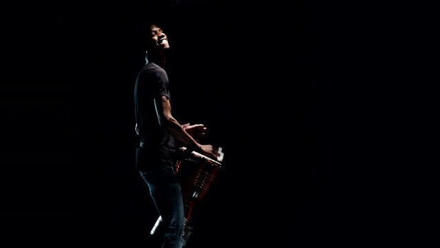 Black musician plays standing the wooden hand drum in a dark studio. Music performer vigorously beating with hands the traditional djembe between his legs