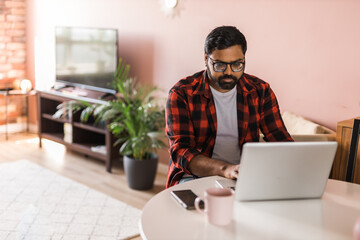 technology remote job and lifestyle concept - happy indian man in glasses with laptop computer working at home office, copy space and empty space for text