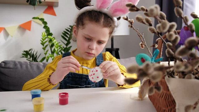Close-up of concentrated cute little girl kid sitting at table painting some eggs for coming Easter. Serious child in fluffy bunny ears playing, drawing and decorating small egg. Spring holiday concep