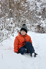 Fototapeta na wymiar A boy in a jacket and a hat sits in the snow and looks into the distance with surprise. Winter vacation.