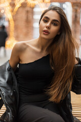 Fashionable beautiful sexy woman with a cute fresh natural face and lips in a trendy black outfit with a one-shoulder top and a down jacket is sitting in the city on a blurred urban background