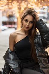 Fashionable beautiful girl model with a sexy one-shoulder top and a down jacket sits on a bench in the city and fixes her hair