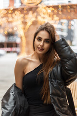 Happy beautiful young woman model in fashion winter clothes with jacket and top sitting on a bench in the city near the vintage lights bokeh