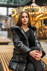 Fashion pretty elegant woman in stylish black winter clothes with down jacket sits on the bench in the city