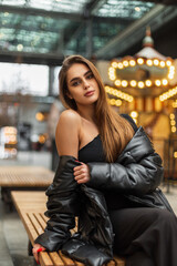 Pretty sexy woman in a fashion black top and a winter down jacket sits on a bench in the city near the vintage carousel with lights