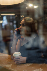 Beautiful girl sits in a cafe near a shop window, drinking coffee and talking on the phone.