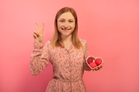 Happy smiling young woman showing peace gesture v sign with two fingers hand and holding heart shaped plate saucer with three dyed painted easter eggs isolated on pink background.

Easter Day concept.