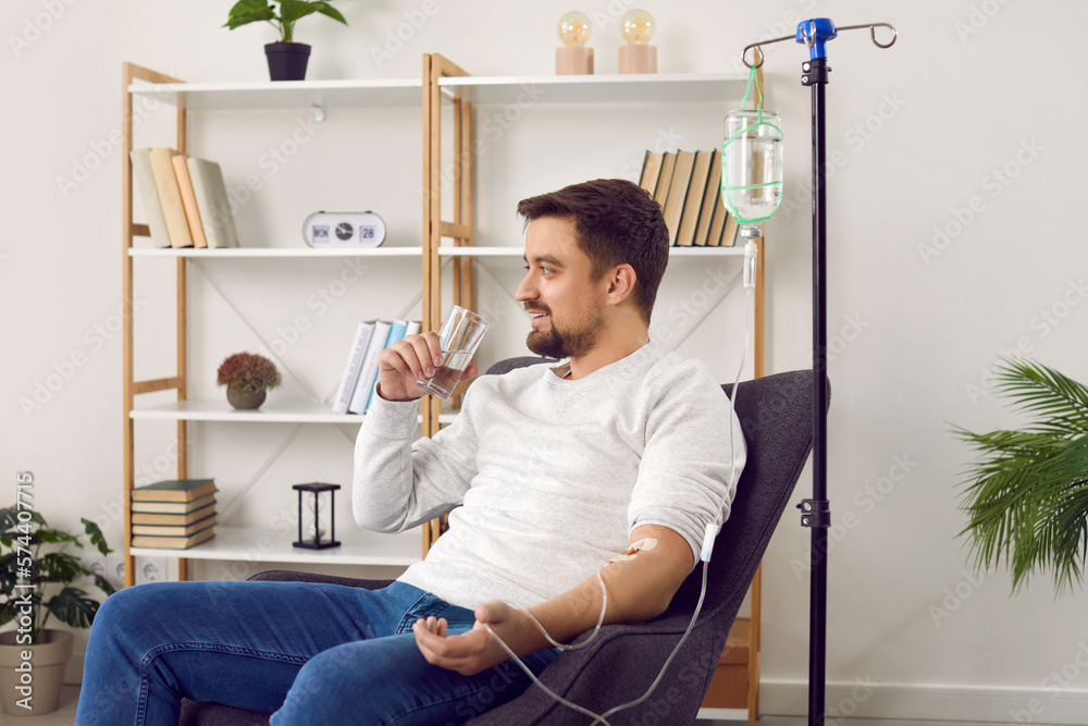 Wall mural Dehydrated young man receiving intravenous vitamin therapy in hospital room. Male patient sitting in armchair attached to vitamin IV infusion drip in wellness center or beauty salon